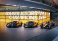 Three black and orange McLaren race cars are displayed indoors, with a large yellow and white "Constructors Champions 2021" sign in the background.