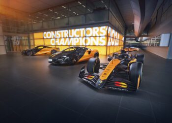 Three sleek orange and black McLaren performance cars, including the 750S and the 2024 MCL38, are parked in a modern garage. A sign in the background proudly reads "CONSTRUCTORS CHAMPIONS," highlighting their engineering excellence.