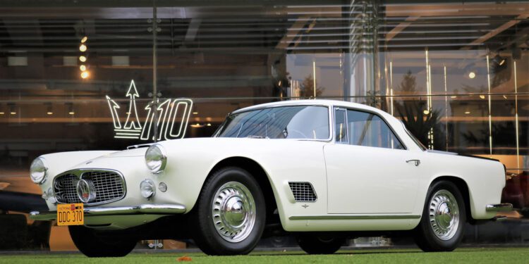 A vintage white Maserati 3500GT is elegantly parked on the green grass in front of a glass building adorned with a "110" logo, showcasing its timeless Classiche charm.
