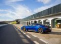 A blue Maserati GT2 Stradale is elegantly parked on a race track near a modern building, set against the backdrop of Spain's partly cloudy sky.