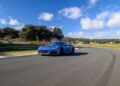 A blue Maserati sports car speeds along a racetrack in Spain, cutting through the clear blue sky adorned with scattered clouds.