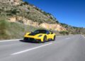 A yellow GT2 Stradale zooms along an open road in Spain, flanked by rocky hills and under a clear blue sky.