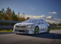 A camouflaged car with green wheels, reminiscent of the BMW Vision design, is driving on a highway surrounded by trees under a partly cloudy sky.
