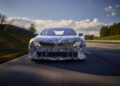 A camouflaged prototype car, reminiscent of the BMW Vision, cruises along a tree-lined road under a cloudy sky. Its distinctive headlights hint at cutting-edge EV tech that could define the future of the Neue Klasse range.