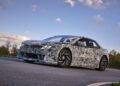 A camouflaged BMW sports car cruises on a paved road under a blue sky with clouds, hinting at the innovative EV Tech of the Neue Klasse.