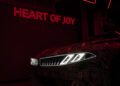 A BMW Vision car with illuminated headlights is partially visible in a dimly lit space. A neon sign reading "HEART OF JOY" glows on the wall behind it, capturing the essence of its advanced EV Tech.