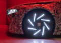 Close-up of a car with a swirling patterned wrap, highlighting bright LED headlights and illuminated wheel spokes in a red-lit environment, showcasing cutting-edge EV tech of the BMW Vision Driving Experience.