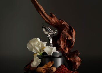 A silver figurine on a round base is surrounded by a white flower, twisted wood, and cinnamon sticks on a dark background, evoking an in-car fragrance as luxurious as the scent within a Rolls-Royce.