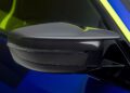 A close-up of a BMW M4 CS VR46's side mirror reveals its sleek carbon fiber finish, adorned with striking blue paint and subtle yellow accents, reminiscent of Valentino Rossi's vibrant style.