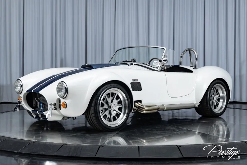 A unique creation, this white vintage-style convertible with blue racing stripes, shiny chrome wheels, and a roll bar is elegantly parked indoors in a custom car garage.