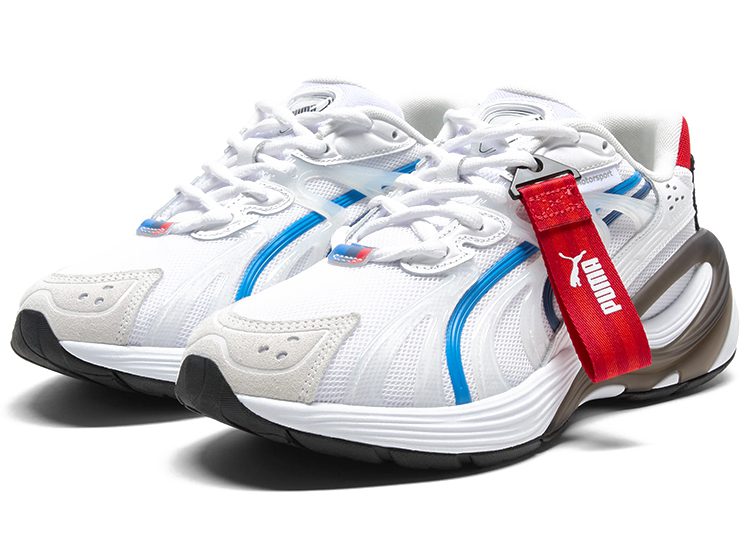 A pair of white PUMA sneakers with blue accents, red heel tabs, and a red Puma-branded strap across the right shoe—an automotive-inspired design for the ultimate driver.