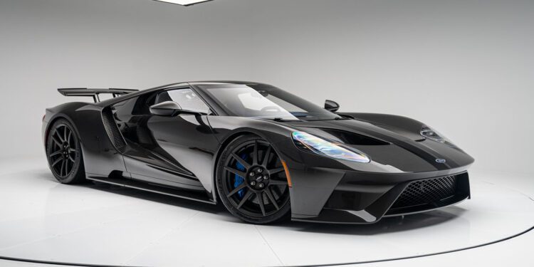 A sleek black Ford GT sports car with a low, aerodynamic design, large wheels, and a rear spoiler is parked in a white studio setting. The Liquid Carbon Edition subtly flaunts ghosted stripes that add an extra layer of intrigue to its flawless finish.
