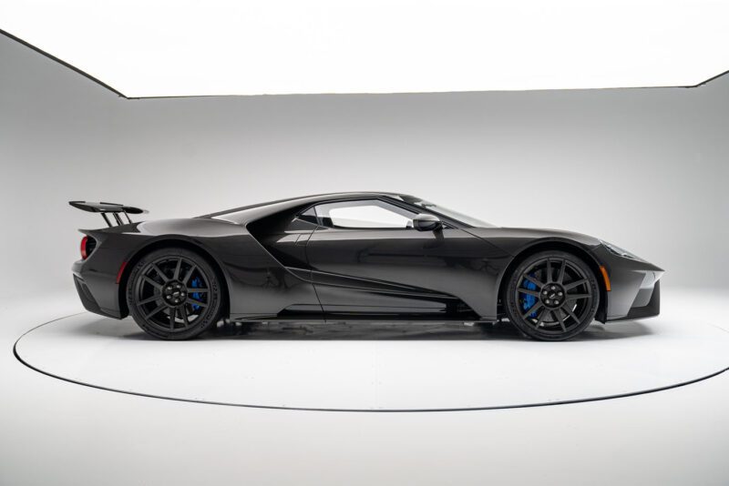 A sleek, black Ford GT Liquid Carbon Edition sports car, featuring ghosted stripes and a large rear spoiler with blue brake calipers, is displayed in a well-lit studio setting.