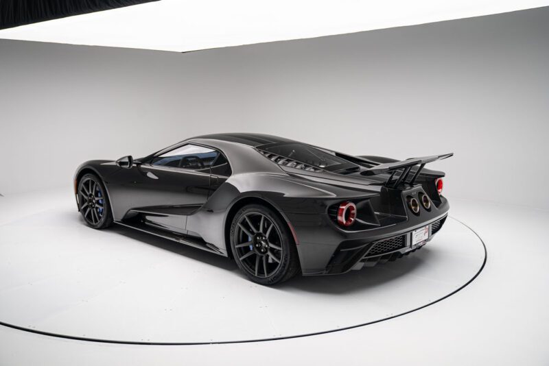 The Ford GT in Liquid Carbon Edition stands proud in a white studio setting, boasting an aerodynamic design with ghosted stripes, a large rear wing, and dual exhausts.