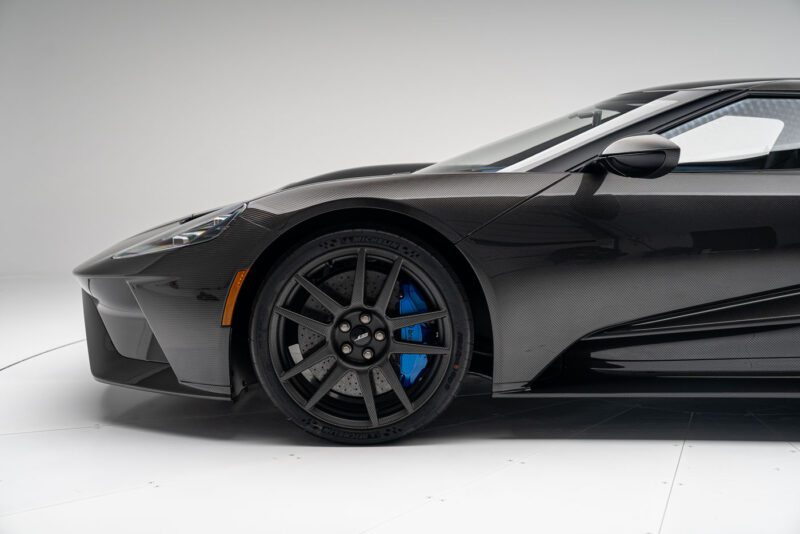 Close-up of a Ford GT's front section in the Liquid Carbon Edition, showcasing its sleek body, detailed wheel with blue brake caliper, ghosted stripes, and aerodynamic design.