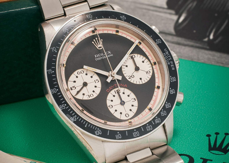 Close-up of a Rolex Cosmograph Daytona watch with a silver case, black bezel, and white subdials, featured in the duPont REGISTRY's Ultimate Weekend Guide. The watch chicly rests on a green surface, embodying luxury and style perfect for ModaMiami 2025.