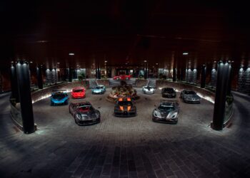 A circular driveway showcases nine parked luxury sports cars, including models in red, blue, silver, and black. The setting is well-lit with columns and spotlights illuminating the scene—a perfect feature for the duPont REGISTRY Supercar Club gathering.