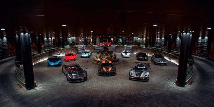 A circular driveway showcases nine parked luxury sports cars, including models in red, blue, silver, and black. The setting is well-lit with columns and spotlights illuminating the scene—a perfect feature for the duPont REGISTRY Supercar Club gathering.