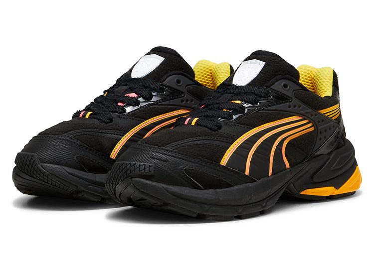 A pair of black PUMA sneakers with yellow accents, orange detailing, and a white logo on the tongue, automotive-inspired and perfect for the driver seeking style and performance, displayed against a white background.