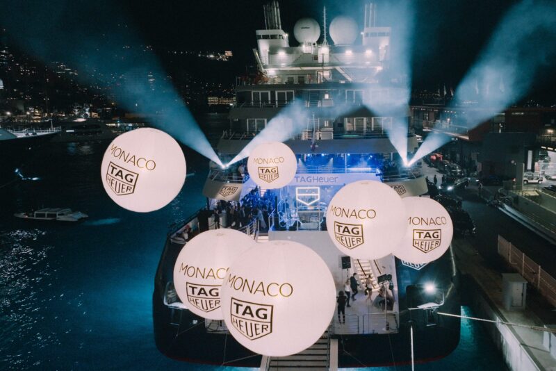 A luxurious nighttime yacht party glowed under beams of light with illuminated balloons showcasing "Monaco TAG Heuer" logos. Guests enjoyed the opulence on deck, reminiscent of the Monaco Grand Prix, celebrating the sophistication of this renowned luxury watch brand.