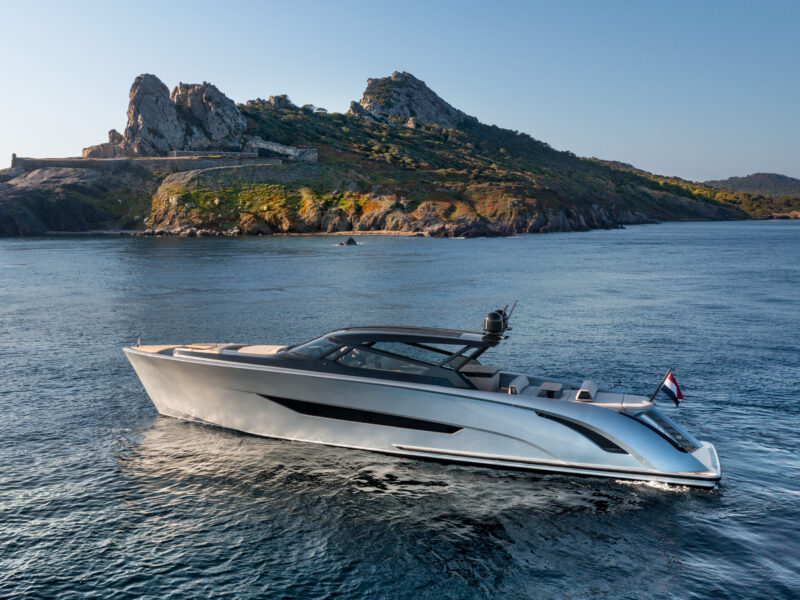 A sleek, modern Wajer Yacht sails in calm waters near a rocky island with lush green vegetation under a clear blue sky, epitomizing innovation and luxury.