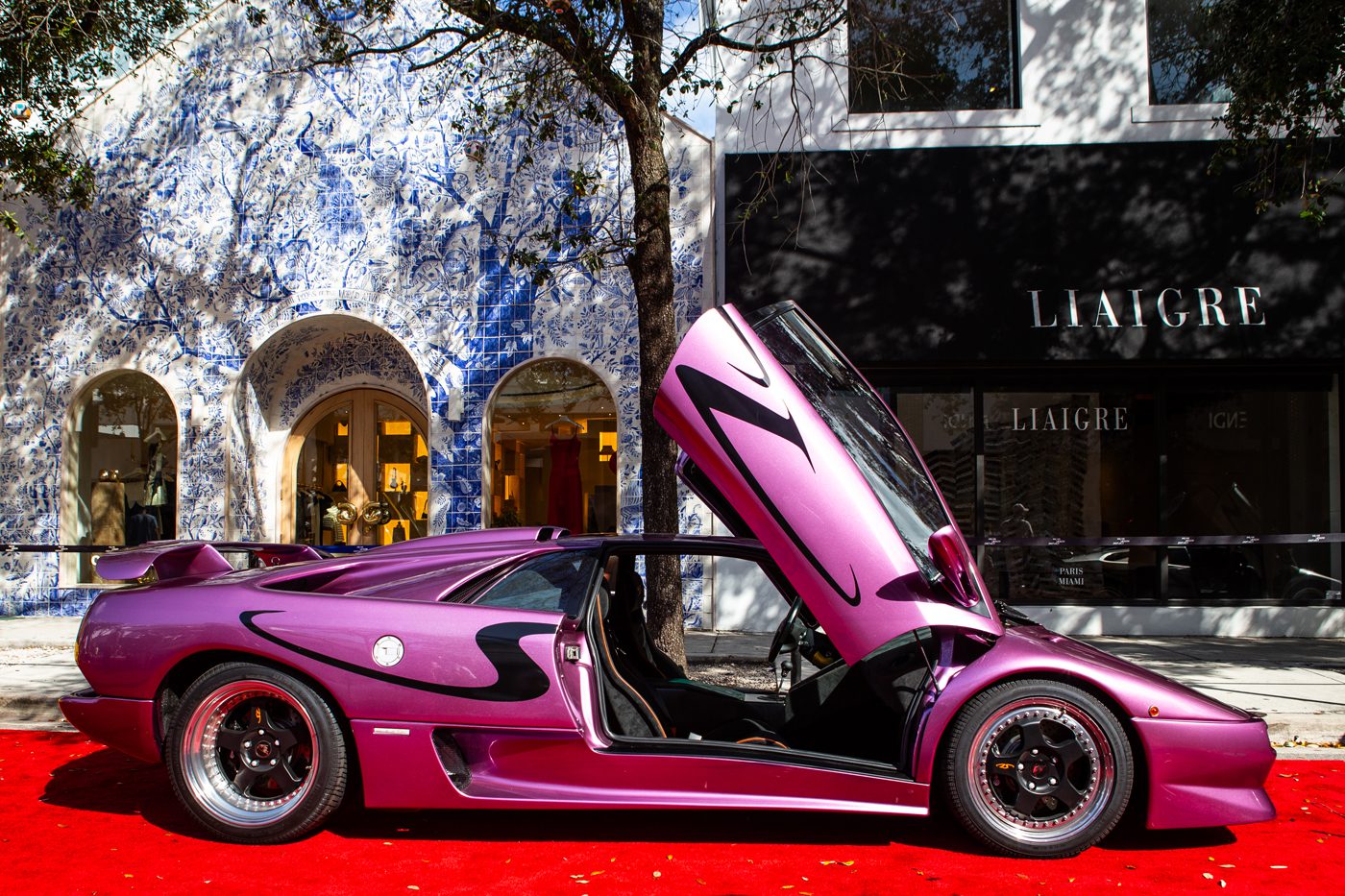 A purple supercar with butterfly doors open stands proudly on a red carpet, set against a backdrop of blue and white wall art by Liaigre. This stunning scene is part of the Miami Concours 2025, where thousands gather to celebrate automotive artistry.