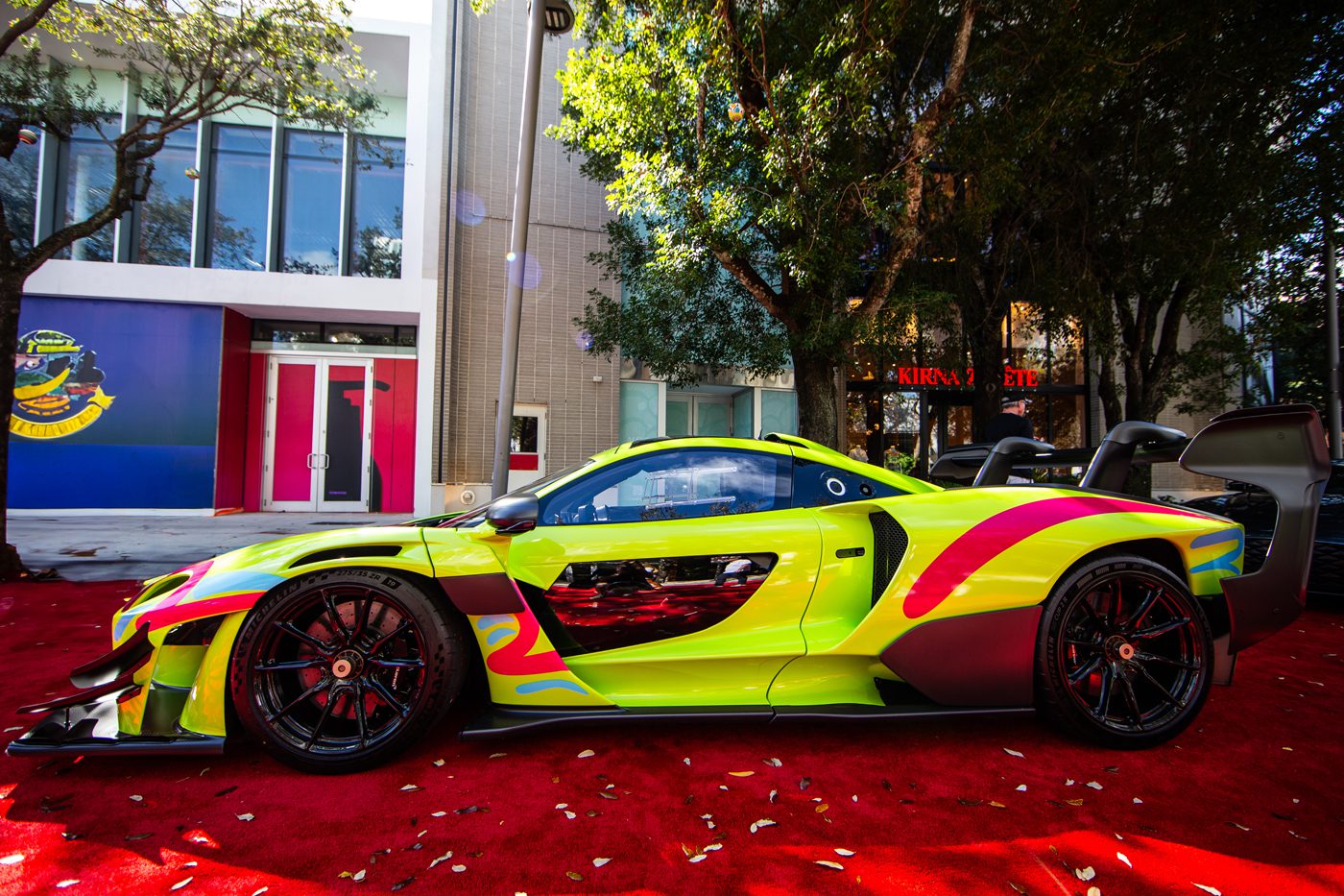 A brightly colored sports car is parked on a red carpet at the Miami Concours, surrounded by trees and modern buildings in the background.