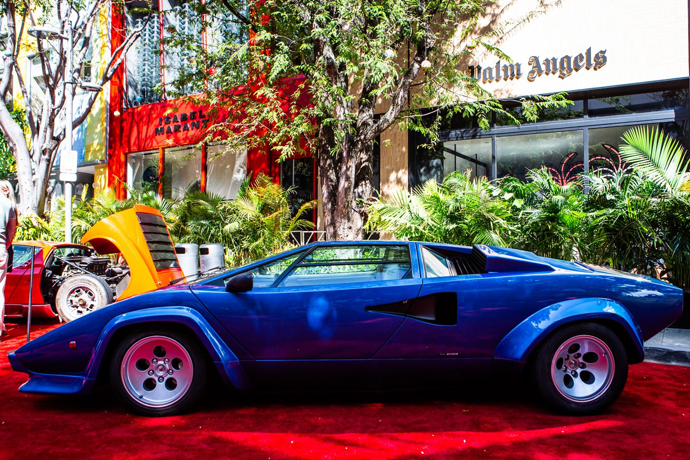 At the Miami Concours 2025, a blue Lamborghini Countach graces the red carpet in front of a store, exuding supercar art. In the background, another car with an open hood adds to the captivating automotive display.