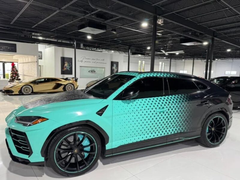A turquoise and black Lamborghini Urus with a geometric dot pattern is parked indoors near a gold Lamborghini in a modern showroom, showcasing one of the best Lamborghini SUVs for sale.