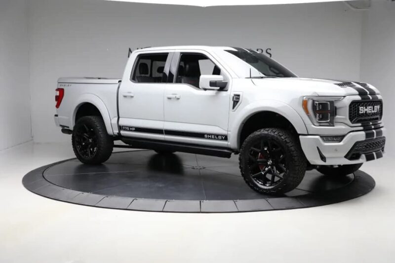 A white Ford F-150 Shelby pickup with black racing stripes, large black wheels, and tinted windows is on display in a studio setting—showcasing the power and style Shelby Trucks are known for. For sale now.