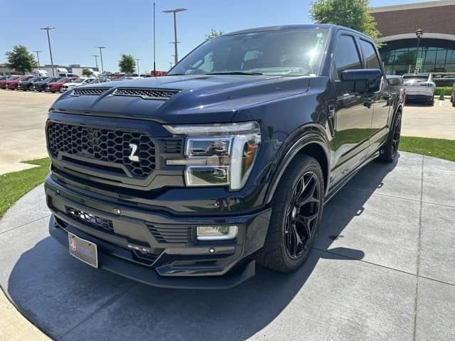 A black Ford F-150 with a custom grille and body kit is parked on a paved surface outside a dealership on a sunny day, showcasing one of the standout Shelby trucks for sale.