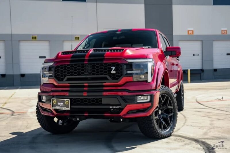 A red and black lifted Ford F-150 with custom wheels is parked in front of a building with several garage doors.