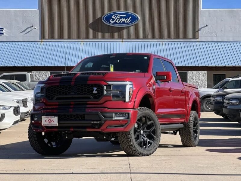 A red Ford F-150 Shelby pickup truck with black racing stripes is parked in front of a Ford dealership, showcasing one of the most eye-catching trucks for sale.