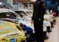 A soccer player in a black jacket stands next to a row of sports cars, including the sleek Porsche Formula E, in a showroom.
