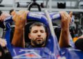 A man sits in a blue race car cockpit, gripping the steering wheel with determination reminiscent of a Porsche Formula E driver, surrounded by people working diligently around him.