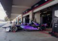 A sleek purple and black Porsche Formula E race car is parked in a Miami garage area, with photographers and crew buzzing nearby. Banners showcasing sponsor names flutter gently, capturing the essence of high-speed excitement akin to a Sergio Agüero goal celebration.