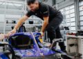 In a garage, a man in a black shirt and pants leans over a Porsche Formula E Car, meticulously adjusting its components.