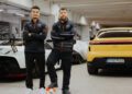 Two men in sports attire stand with arms crossed in a garage. One rests a foot on a soccer ball, embodying the poise of a soccer player between a white car on the left and a yellow Porsche Formula E on the right.