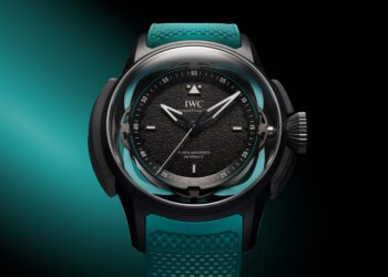 A black watch with a textured dial, luminescent hands, and teal accents is elegantly showcased against a gradient aqua and black background, inspired by the Toto Wolff Edition Big Pilot’s Watch.