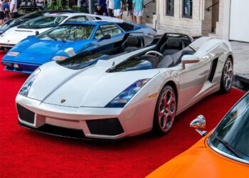 A white convertible sports car gleams on the red carpet amidst other luxury vehicles at the Miami Concours. Elegantly dressed attendees linger in the background beside Moore Private Members’ Club, creating an air of exclusivity and allure.