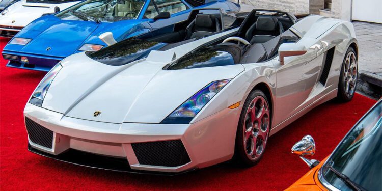 A white convertible sports car gleams on the red carpet amidst other luxury vehicles at the Miami Concours. Elegantly dressed attendees linger in the background beside Moore Private Members’ Club, creating an air of exclusivity and allure.