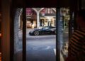 A sleek black Rimac sports car is parked on a street lined with luxury storefronts, making its debut appearance. In the foreground, a person in a striped sweater is partially visible from inside a building.