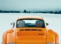 A unique display of power and elegance, this orange sports car flaunts its "Rodeo" badge proudly as it cruises down a snow-covered road. Captured from the rear with its license plate and spoiler in view, the scene is framed by the ethereal clouds of I.C.E. St. Moritz.