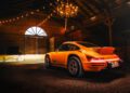 An orange sports car, reminiscent of a RUF Rodeo, is parked in a dimly lit wooden barn with string lights and a chandelier hanging from the ceiling.