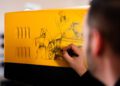 A person, inspired by the elegance of a Bugatti Baby II, sketches a horse and a machine on a yellow and black surface with precision, capturing the spirit of the masterpiece.