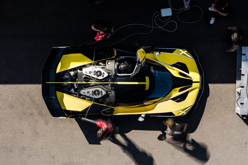 Pemandangan overhead dari hypercar kuning dan hitam di area pit stop, anggota kru sibuk. Kokpit mobil terbuka, peralatan tersebar di dekatnya, siap untuk tes penggeledahan berikutnya.