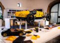 A yellow vintage-style car, reminiscent of the Bugatti Baby II, stands proudly on a display table in an exhibition room. Around it, Hedley Studios showcases design materials and miniature models on the foreground table.