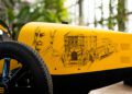 Close-up of a vintage yellow car's side, featuring sketches of a man, a historic building, and classic cars with handwritten text from Hedley Studios. Among the designs is a nod to the Bugatti Baby II, capturing timeless elegance in automotive artistry.