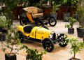 A small, yellow vintage-style toy car, reminiscent of a Bugatti Baby II, is parked on a tiled floor surrounded by potted plants. Just behind it, crafted by Hedley Studios, stands a black antique-style tricycle with a brown seat.