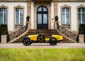 A yellow vintage Bugatti Baby II is parked in front of a grand building with arched windows and ornate railings, reminiscent of a classic scene captured by Hedley Studios.